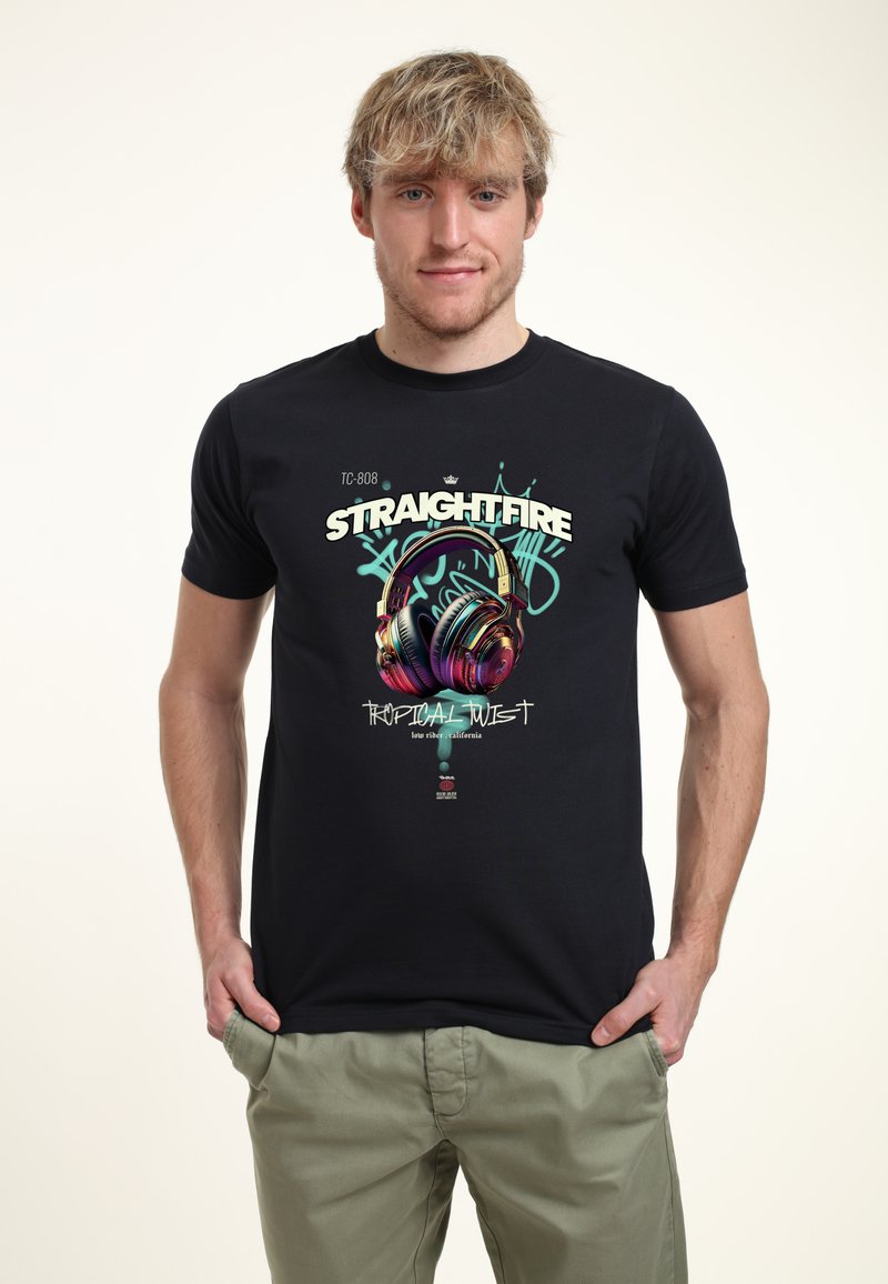 Černé bavlněné tričko s grafickým potiskem, které zobrazuje sluchátka a text "STRAIGHTFIRE TROPICAL TWIST." Model má na sobě khaki kalhoty.