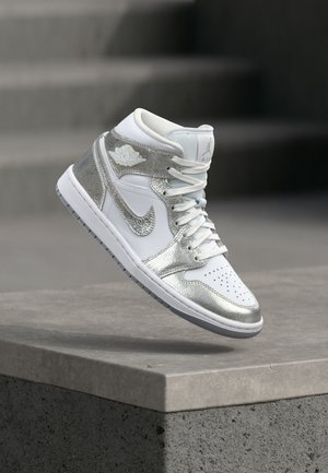 Valge ja hõbedane Nike Air Jordani kõrge säärega toss, mis hõljub betoonserva kohal, taustal on uduselt näha trepiastmed.