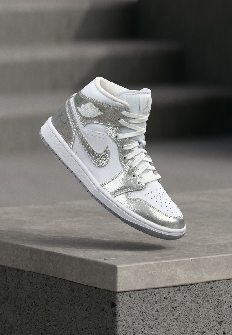 Hvit og sølvfarget Nike Air Jordan høyt sneakers svever over en betongkant med uskarpe trinn i bakgrunnen.