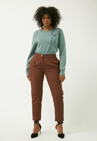 Maglione lungo teal a maniche lunghe con dettagli in perle e cinturino, abbinato a pantaloni beige eleganti a vita alta e scarpe nere con tacco.