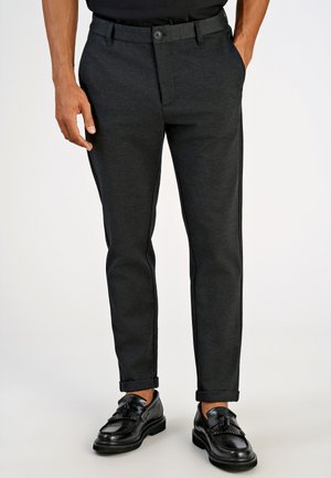 Mand iført mørkegrå slim fit bukser med opkavsede manchetter og sorte læder tassel loafers, der står mod en ensfarvet baggrund.