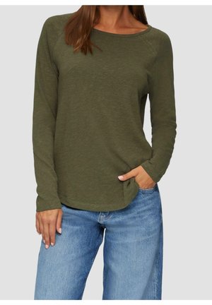 Frau trägt olivgrünes Longsleeve mit Bootsausschnitt und hellblaue Jeans mit hoher Taille, eine Hand in der Tasche, vor neutralem Hintergrund.