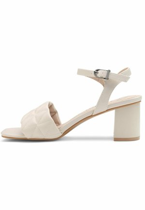 Sandale à talon beige avec un design de lanière matelassée, bride de cheville réglable avec boucle en métal et talon bloc pour plus de stabilité.