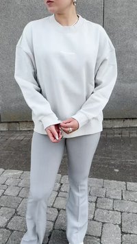 Világosszürke, túlméretezett pulóver, bordázott mandzsettákkal és alsó résszel, amelyen diszkrét logó található a mellrészen. Hozzá illő, testhez simuló világosszürke nadrággal párosítva.