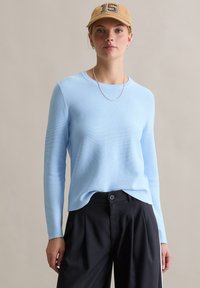 Lichtblauwe gebreide trui met lange mouwen en een geribbelde structuur, gecombineerd met een zwarte broek en een beige pet met een nummer.