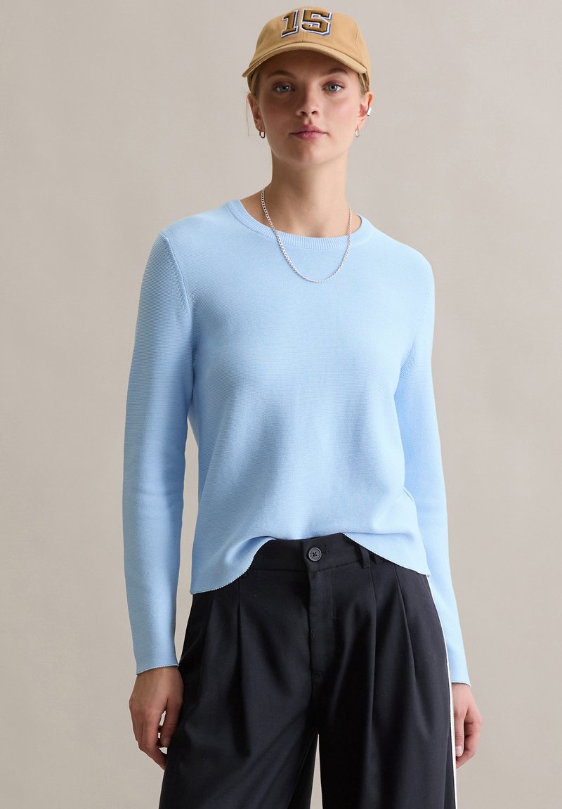 Lichtblauwe gebreide trui met lange mouwen en een geribbelde structuur, gecombineerd met een zwarte broek en een beige pet met een nummer.