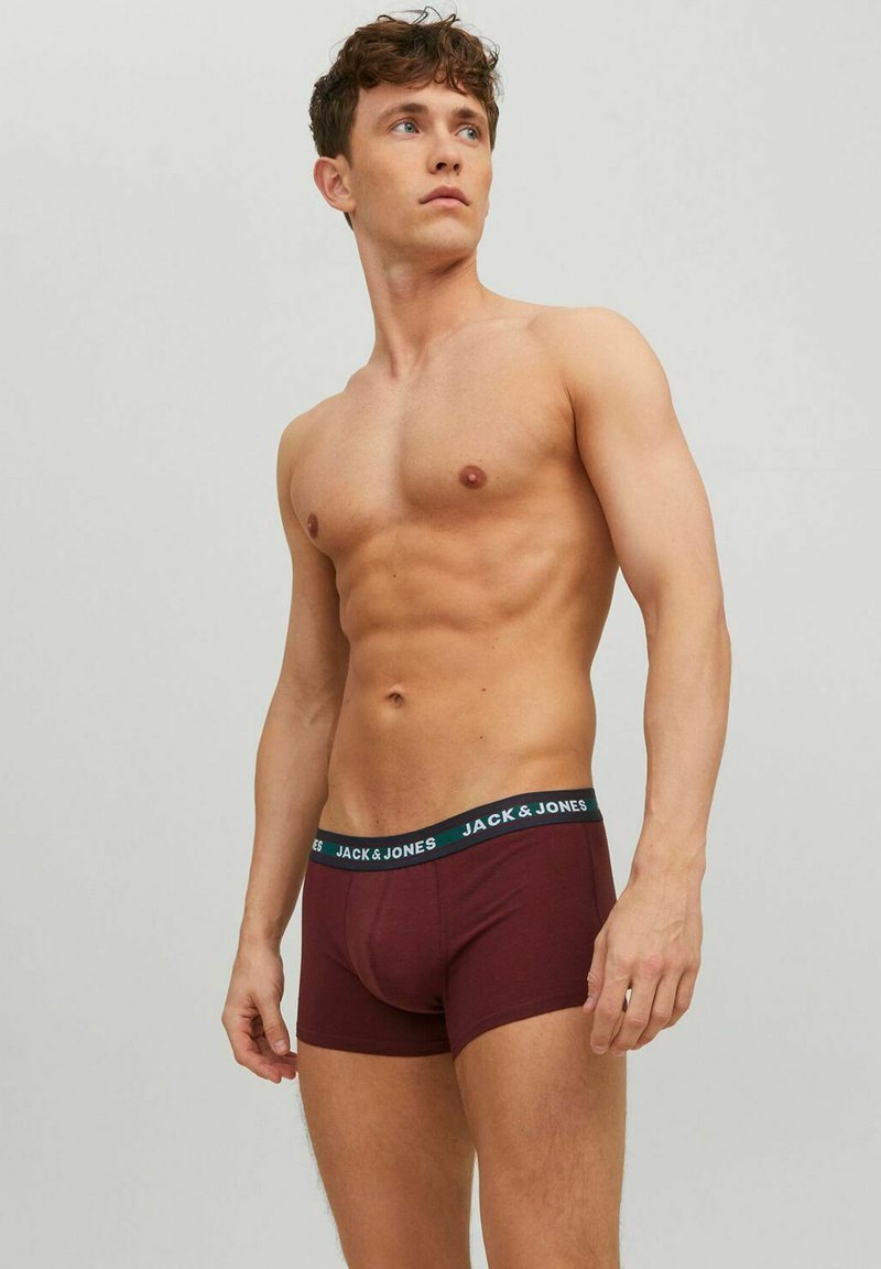 Young muscular man wearing maroon Jack & Jones boxer briefs, looking to the side, standing against a plain light gray background.
