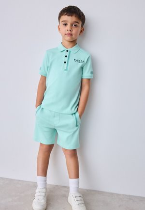 Poiss kannab hele sinist polosärki ja sobivaid lühikeseid pükse, seisab käed taskutes vast seina ääres ja valged tennised jalas.