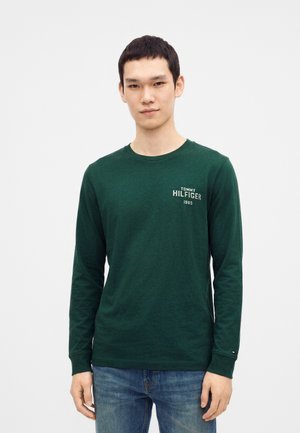Jeune homme portant une chemise à manches longues Tommy Hilfiger vert foncé et un jean bleu, se tenant devant un fond blanc uni.