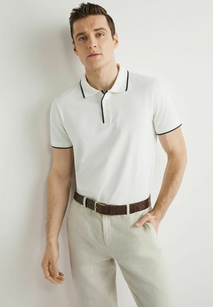 Młody mężczyzna w białej koszulce polo z czarnym obszyciem, beżowych spodniach i plecionym brązowym pasku stoi na tle białej ściany.