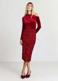 Haut en velours rouge à épaules dénudées associé à une jupe midi ajustée en sequins rouge assorti. La tenue est complétée par des chaussures à talons rouges ornées de nœuds.