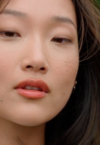 Primer plano de la cara de una mujer con piel suave, maquillaje natural, labios llenos en un tono de coral suave y cabello oscuro que enmarca parcialmente su rostro.