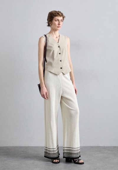 Beige mouwloos vest met ronde zwarte knopen, gecombineerd met witte geribde wijde broeken met een zwart patroon aan de randen. Zwarte sandalen.