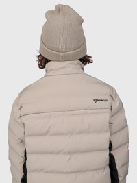 Beige pufi dzsekit viselő személy, rajta a "Brunotti" logóval, és egy színben illeszkedő bézs kötött sapkával, hátulról nézve.
