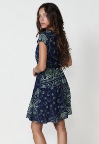 Rochie bleumarin cu un model paisley verde, mâneci scurte, bust ajustat și fustă evazată. Material neted, lungime până la genunchi, croi obișnuit, fără închideri vizibile.