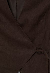 Tmavě hnědá látka ve stylu zavinovacího oděvu s detailem uvázaného uzlu, zobrazující texturu a šití na oblečení zblízka.