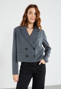 En kvinne med skulderlangt bølgete hår har på seg en grå dobbelspent blazer og svarte bukser, og poserer mot en ensfarget hvit bakgrunn.