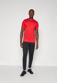 Rød atletisk skjorte med korte ærmer og sorte detaljer; parret med sorte joggerbukser med et lille logo. Model iført sorte sko.