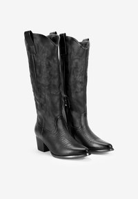 Botas de vaquero de cuero negro con puntera afilada, patrones grabados en la parte superior y un tacón ancho. Presentan una cremallera lateral y una construcción robusta.
