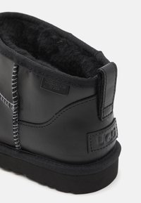 Botín negro de cuero hasta el tobillo con forro de lana, detalles de costura, suela de goma suave y parche con el logo de UGG en el talón.