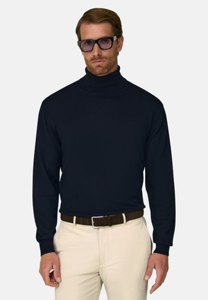 Man draagt een zwarte coltrui, beige broek met een bruine riem en donkere rechthoekige bril, staat tegen een effen achtergrond.