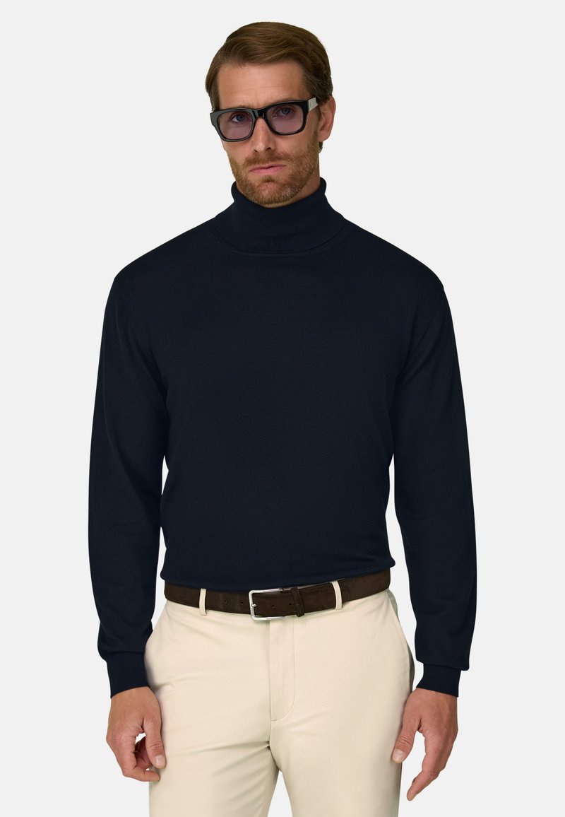 Homme portant un pull à col roulé noir, un pantalon beige avec une ceinture marron et des lunettes rectangulaires foncées, debout devant un fond uni.