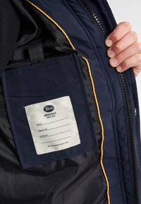 Interno di una giacca navy con un'etichetta su fondo bianco, bordi arancioni e una tasca interna sicura per riporre oggetti.