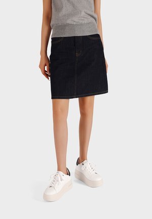 Persona che indossa una gonna di jean scura, una maglietta senza maniche grigia e sneakers bianche con plateau, in piedi contro uno sfondo chiaro e semplice.
