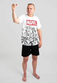 T-shirt bianca con logo rosso "MARVEL" e grafica di supereroe in bianco e nero; pantaloni corti neri; a piedi nudi, in piedi con una mano alzata.
