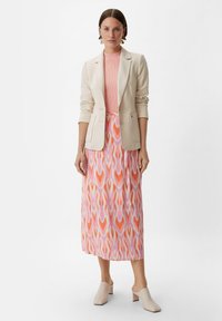 Beige Blazer über einem gerippten rosa Top, kombiniert mit einem langen Rock mit einem mehrfarbigen, abstrakten Herzmuster und hellen Blockabsatz-Mules.