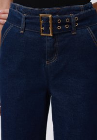 Tumši zilas džinsa bikses ar oranžiem diegiem un plašu jostu, kas apļo ar taisnstūra misiņa sprādzi un vairākiem caurumiem.