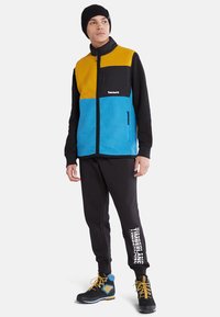 Fargeblokket fleecejakke med gule, svarte og blå paneler, med glidelås og svart logo. Kombinert med svarte joggebukser og støvler.
