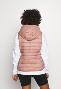 Różowa pikowana kamizelka puchowa z kapturem i poziomymi przeszyciami. Nałożona na biały sweatshirt i w połączeniu z czarnymi szortami.