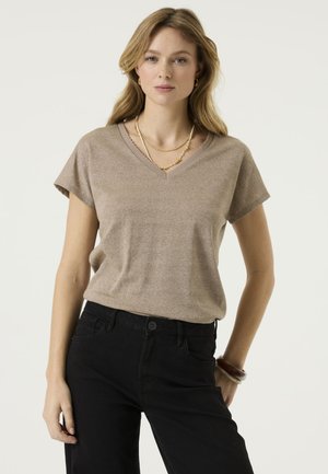 Femme portant un haut beige à manches courtes en V, un pantalon noir, des colliers superposés et des bracelets, debout devant un fond clair uni.