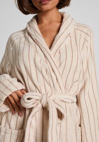 Beżowy szlafrok z jasnobrązowymi pionowymi paskami, wykonany z teksturowanej tkaniny. Posiada kołnierz szalowy, pasek do wiązania oraz przednie kieszenie.