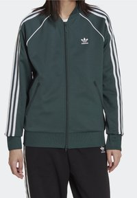Green zip-up track jacket with white stripes along the arms, ribbed cuffs and hem, featuring an embroidered Adidas logo on the chest.