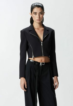 Chaqueta negra corta con mangas largas, cierre de cremallera plateado y solapas puntiagudas. Lleva encima de pantalones negros de tiro alto con un cinturón brillante.