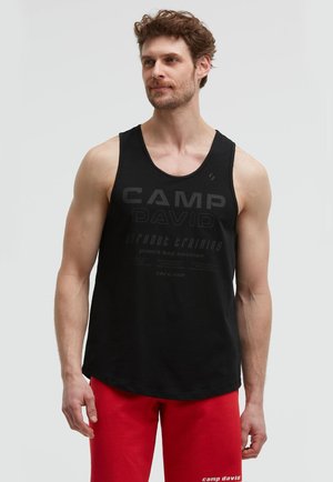 Man with light skin and curly brown hair wearing a black sleeveless gym shirt and red shorts against a plain light background.