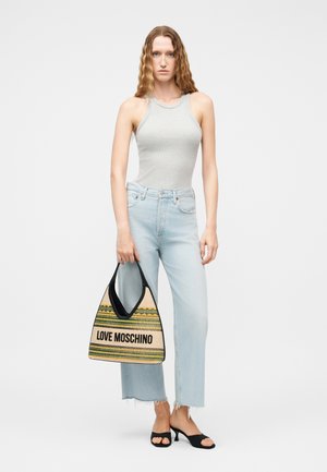 Γυναίκα που στέκεται, φοράει γκρι αμάνικη μπλούζα, ανοιχτόχρωμα τζιν παντελόνια cropped, μαύρα πέδιλα με τακούνι, κρατώντας μια μεγάλη υφαντή τσάντα Love Moschino.