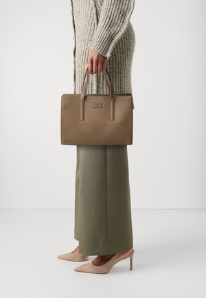 Person in beige knit sweater and olive skirt holding a taupe rectangular handbag, wearing beige high-heeled shoes on white background.
