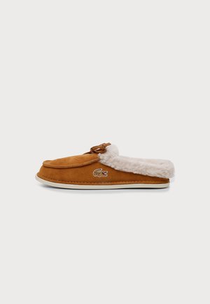 Brun ruskind slip-on tøfler med hvid blød foring og et lille broderet krokodillelogo på siden, sat mod en ensfarvet baggrund.