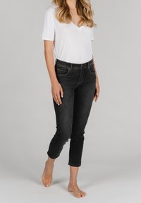 Jeans de mezclilla negra con un corte ajustado y longitud cropped, combinados con una camiseta blanca de manga corta y escote en V. Modelo descalzo se encuentra de pie contra un fondo neutral.
