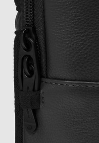 Zwarte leren tas met een textuur, voorzien van een soepele ritssluiting, contrasterende nylon accenten en een webbingriem voor een veilige sluiting.