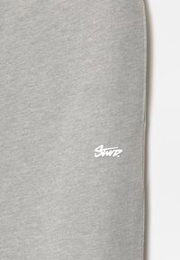 Détail en tissu gris clair avec petit logo blanc stylisé "Swr." imprimé près du bord, fond uni, texture lisse visible.