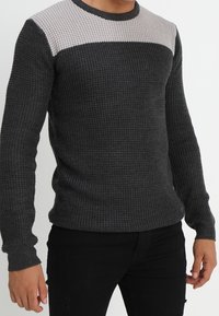 Stickad tröja i mörkgrått med ljusgrå texturerad yoke, rundhals, och långa ärmar. Materialet verkar varmt och något elastiskt.