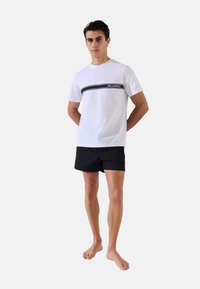 T-shirt bianca in cotone con una striscia orizzontale nera e logo sul petto, abbinata a pantaloni corti neri, modello in piedi a piedi nudi.