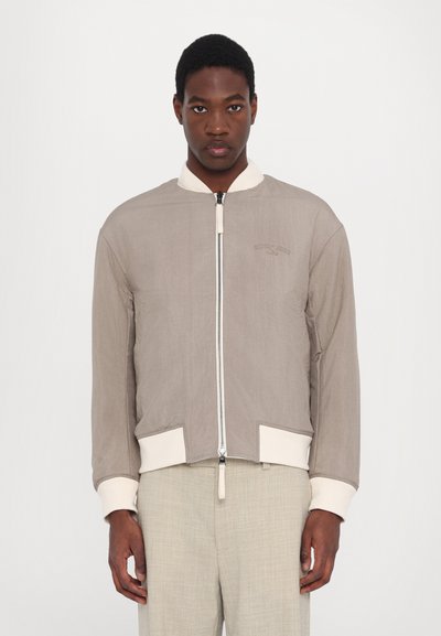 Veste bomber beige clair avec fermeture éclair sur le devant, tissu texturé, poignets et col crème. Comprend deux poches avant angulaires.