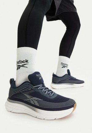 Gambe con leggings neri e calzini bianchi Reebok che indossano scarpe da corsa Reebok blu navy con suole imbottite bianche su sfondo bianco.