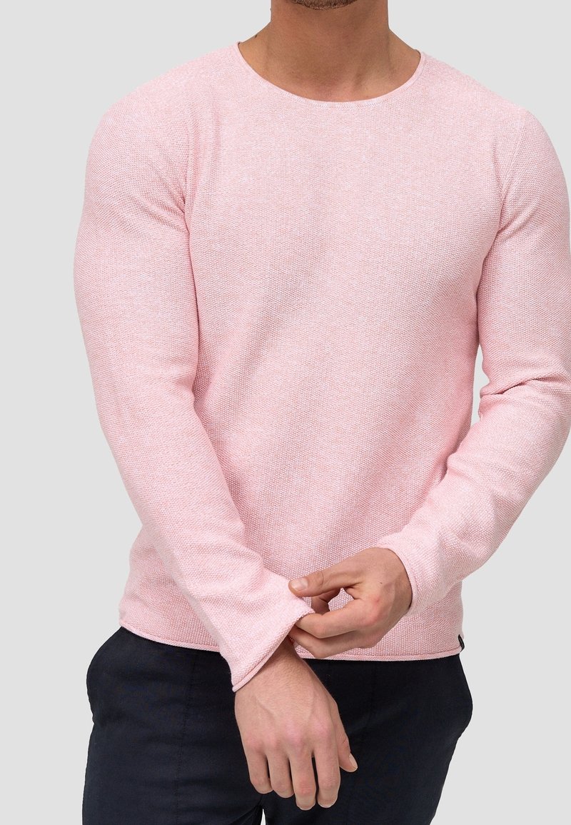 Homme ajustant le poignet d’un pull à manches longues rose clair, portant un pantalon sombre, sur un fond gris clair uni.