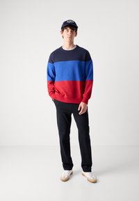 Color-blocked sweatshirt in navy blue, sky blue, and red. Casual fit, round neck, and ribbed hem. Paired with black pants and white sneakers.